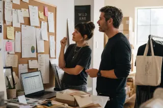 Boutique owner and digital consultant examining business notes and analytics in a sunlit shop backroom, suggesting transition from retail to e-commerce.